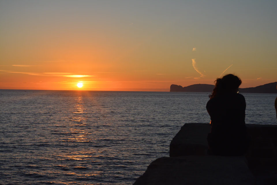 Tramonto sul mare con la sagoma del promontorio di Capo Caccia e una figura seduta sugli scogli