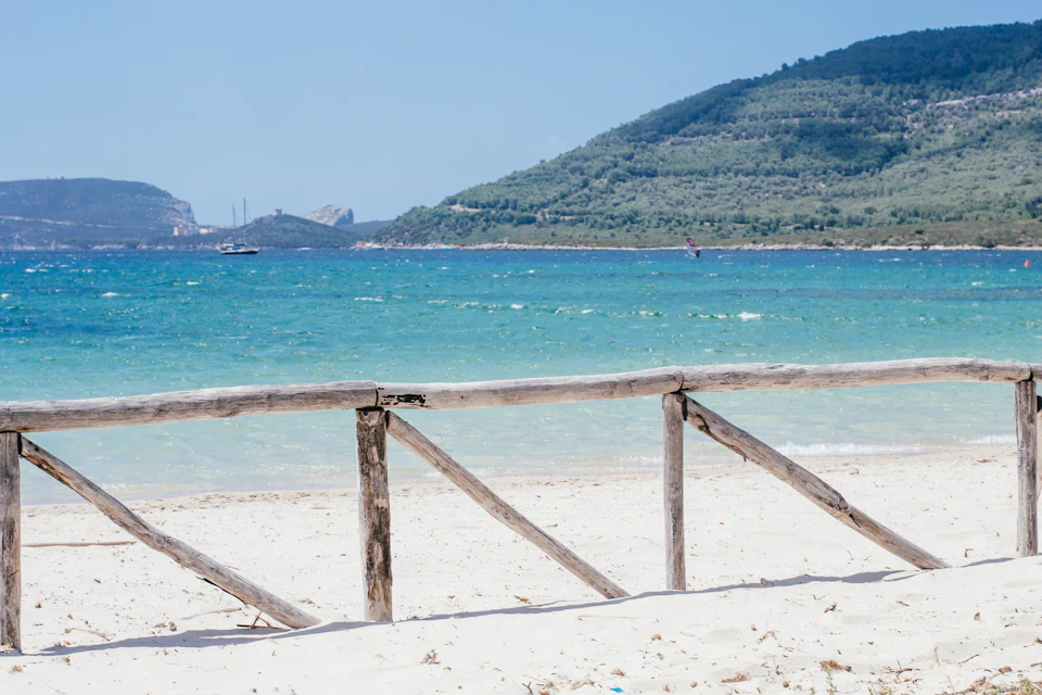 Spiaggia bianca con staccionata di legno, acqua turchese e promontorio verde sullo sfondo